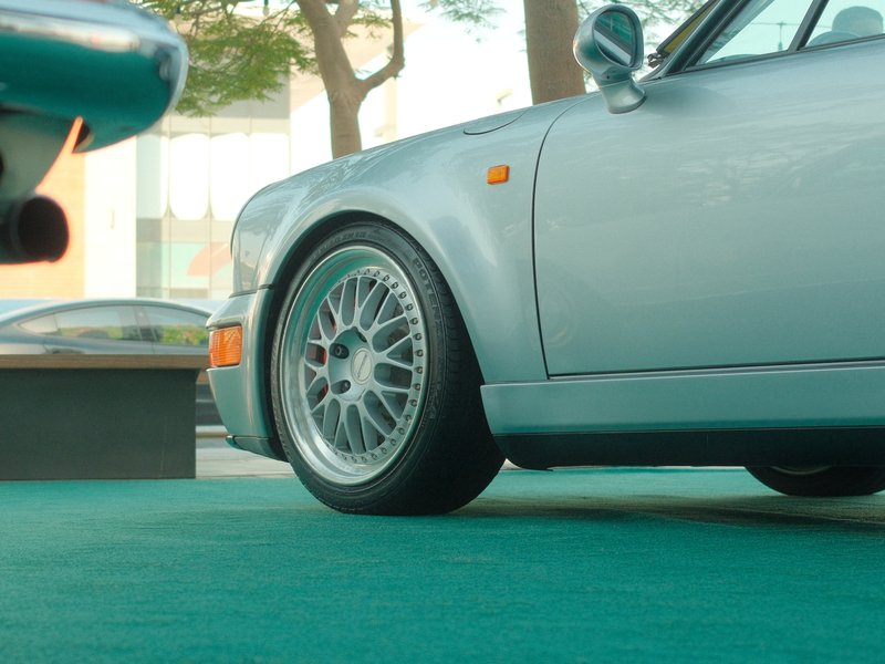 Close-up of a silver car's wheel and fender on a green surface, with blurred background elements.