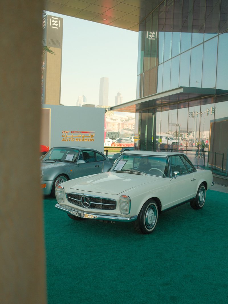 A classic white Mercedes-Benz parked in front of a modern glass building with a city skyline in the background.