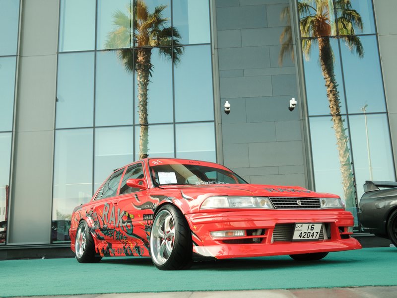 A modified red car with custom graphics parked in front of a glass building and palm trees.