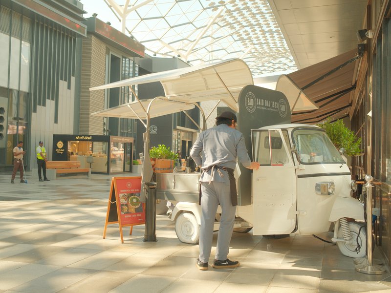 A man at a food stall in a modern urban setting with geometric architecture and bright sunlight.