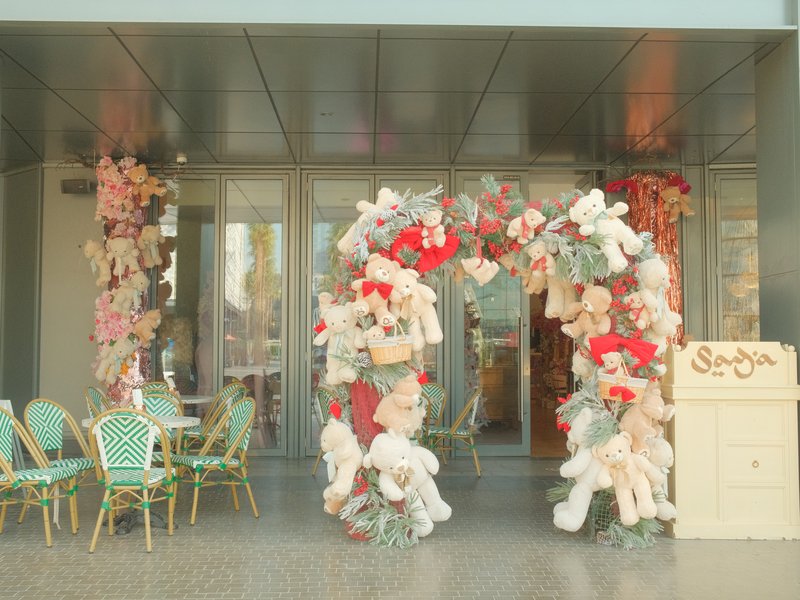 A floral archway adorned with pink and red flowers at a cafe entrance, with green chairs nearby.
