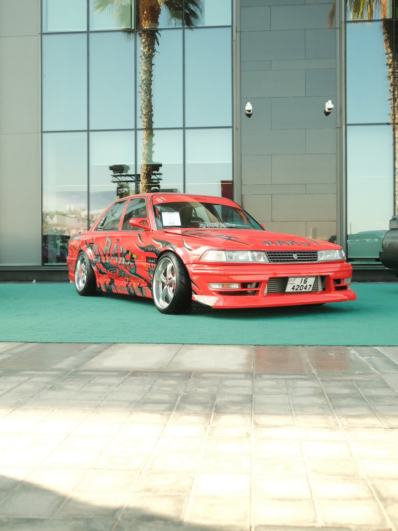 A modified red car with custom graphics parked in front of a glass building, palm trees in the background.