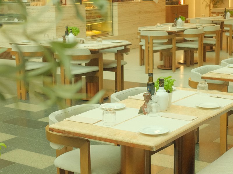 A cozy restaurant interior with wooden tables and chairs, set for dining with white plates and green plants.
