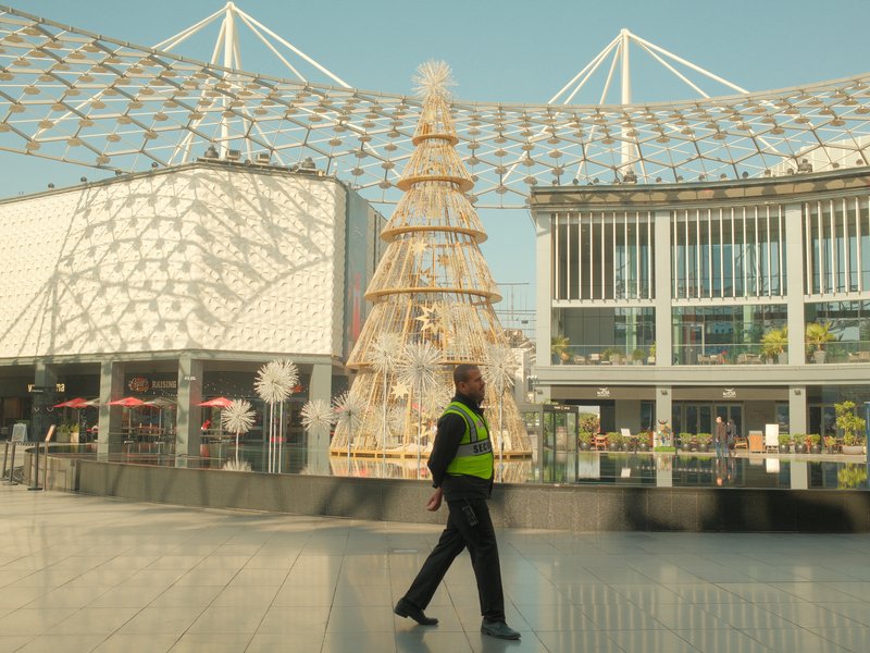 A security guard walking past a large decorative Christmas tree in a modern urban plaza.