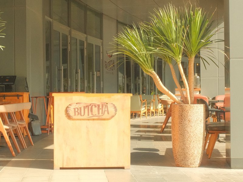 A wooden sign reading 'Butcher' in front of a modern restaurant with a large potted plant.