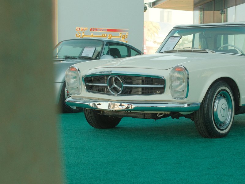 A classic white Mercedes-Benz car displayed at an event, with a green carpet and another car in the background.