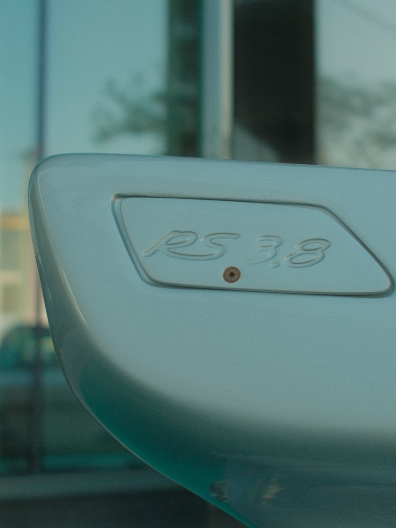 Close-up of a car's rear wing with the model number RS 3.8 engraved on it, reflecting a modern building.