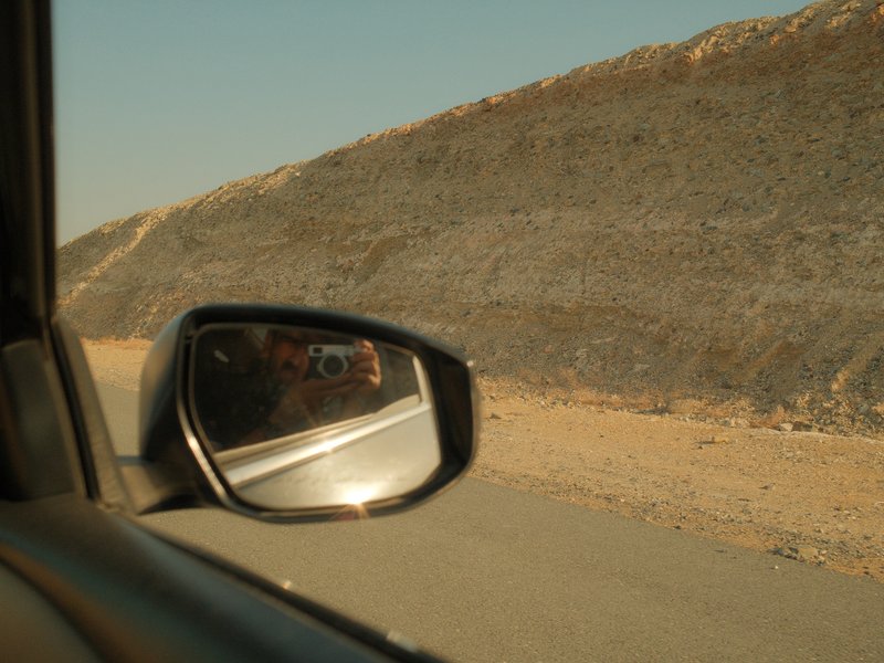 Side view mirror reflecting a rocky hillside along a road under a clear sky.