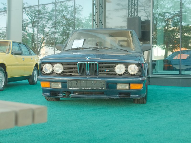 A classic blue BMW car displayed indoors with a green carpet and other vintage cars in the background.