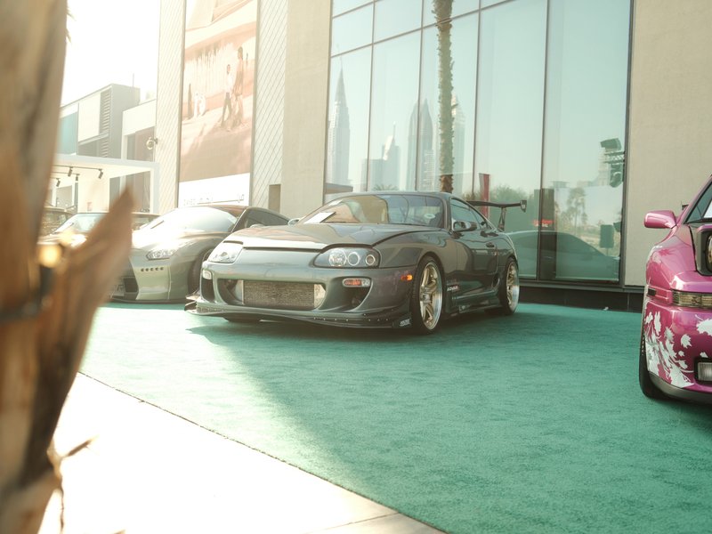 A lineup of modified cars parked on a green carpet outside a modern building with large windows.
