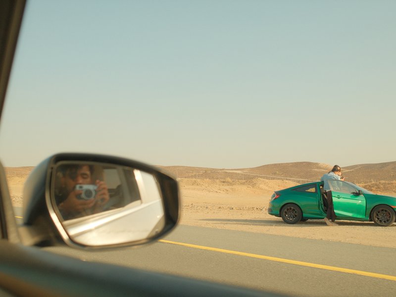 A green car parked on a desert road with a person standing beside it, reflected in a side mirror.