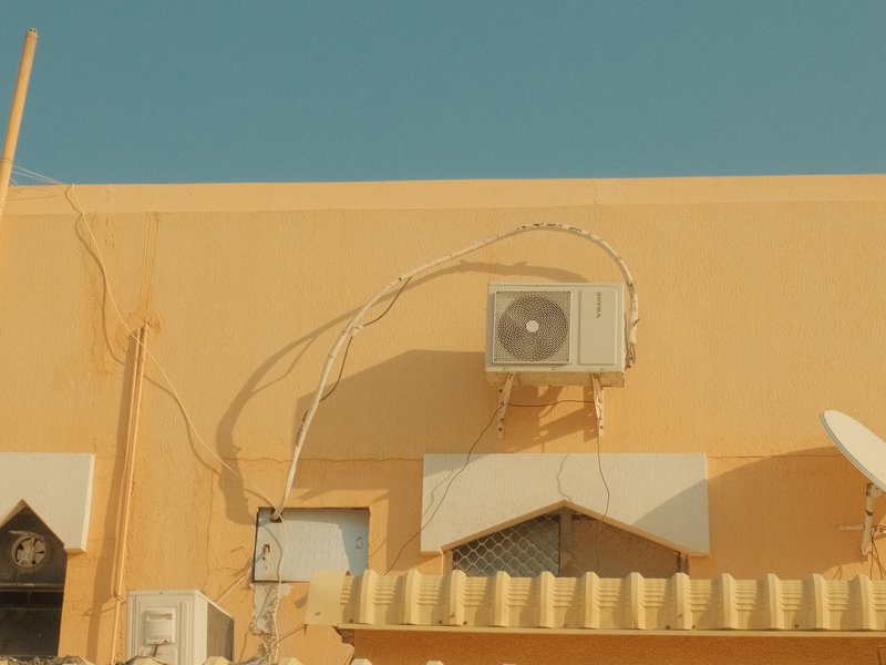An air conditioning unit mounted on a yellow wall with visible wires and a clear blue sky.