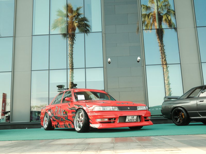 A modified red car with racing decals parked in front of a glass building, palm trees in the background.