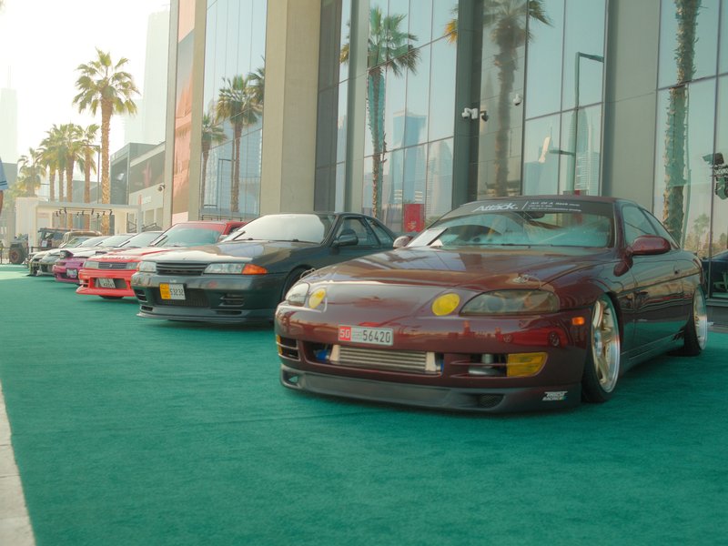 A row of modified cars displayed on a green carpet outside a modern building with palm trees.