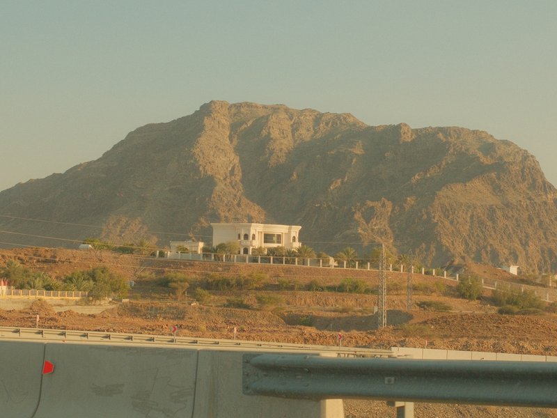 A white house on a hillside with a rocky mountain backdrop under a warm sky.