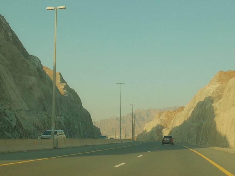 A highway cutting through rocky mountains under a clear sky, with vehicles in the distance.