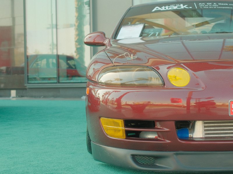 Close-up of a maroon sports car with a yellow fog light, set against a modern building backdrop.