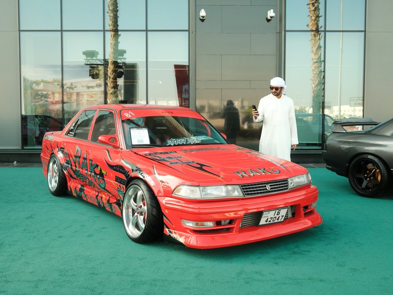 A red modified car with graphics parked on a green carpet, with a man in traditional attire standing nearby.