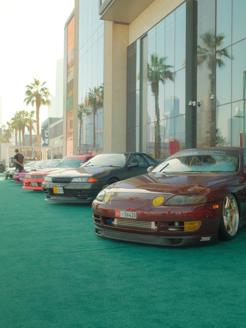 A row of modified cars parked on a green carpet outside a modern building with palm trees.