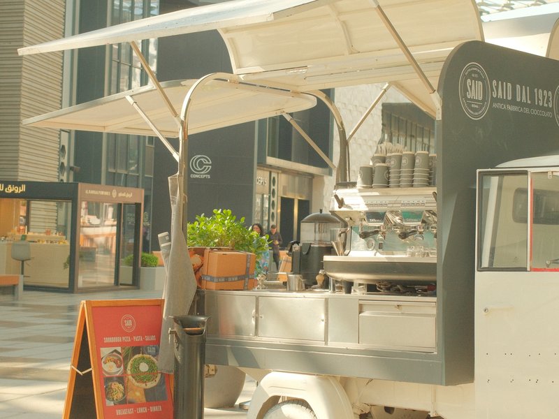 A food truck with an awning parked in an urban area, surrounded by shops and greenery.