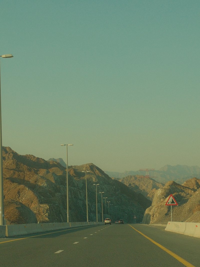 A winding road through rocky terrain under a clear sky, with distant mountains visible.
