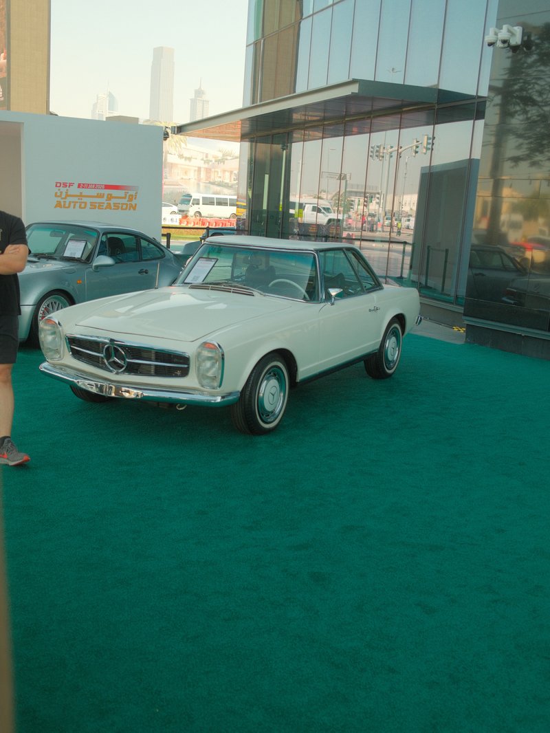 A classic white Mercedes-Benz car parked on green carpet outside a modern building.