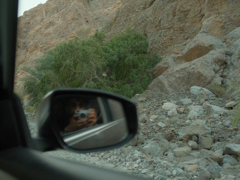 A car side mirror reflecting a person and rocky terrain in the background.