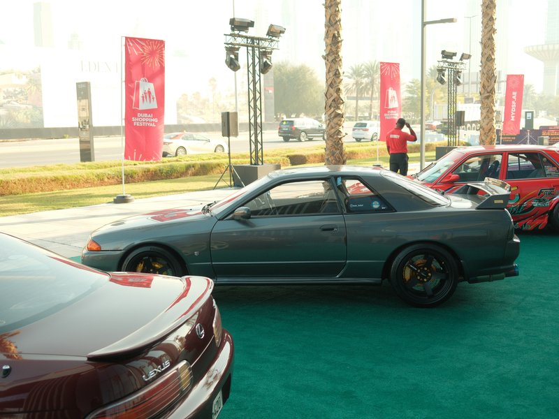 A gray sports car parked at an event with palm trees and banners in the background.
