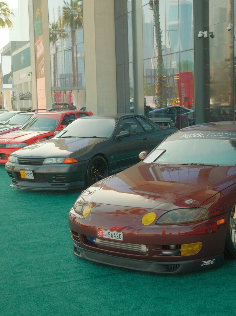 A row of modified sports cars parked on green carpet outside a building, showcasing their vibrant colors.