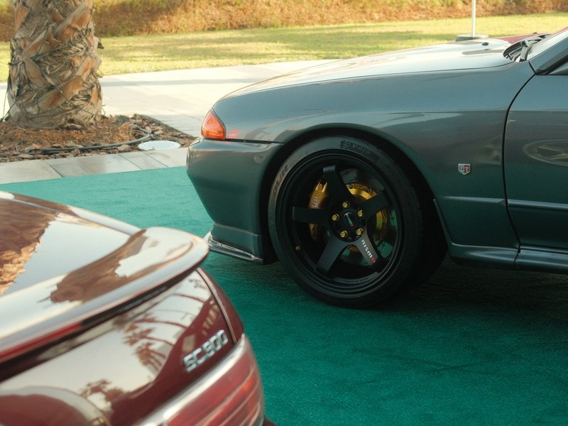 Close-up of two sports cars parked on green carpet, showcasing wheels and body details.