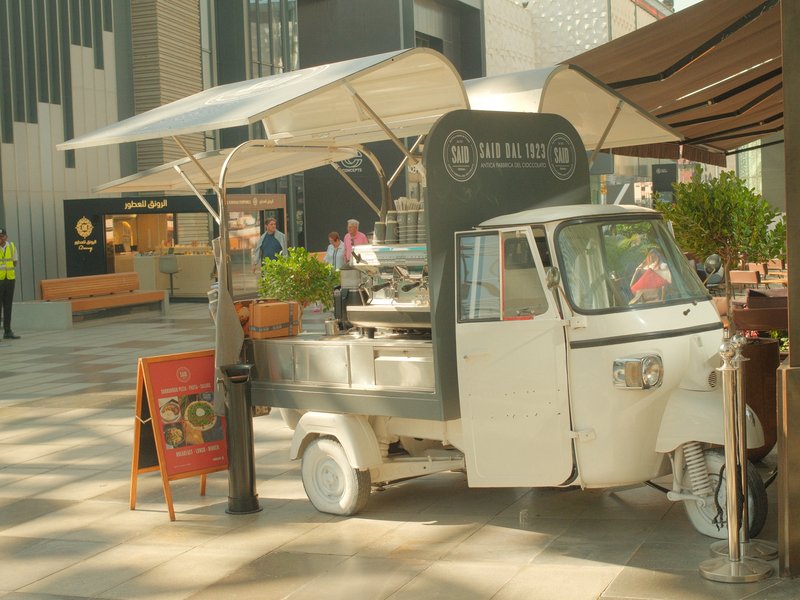 A white food truck with an awning parked in a modern urban area, surrounded by greenery and buildings.