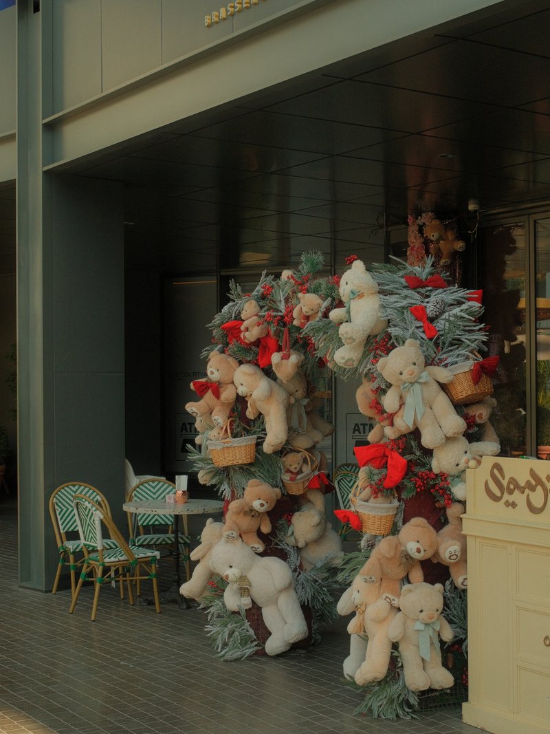 A festive display of teddy bears adorned with red bows and surrounded by greenery outside a cafe.