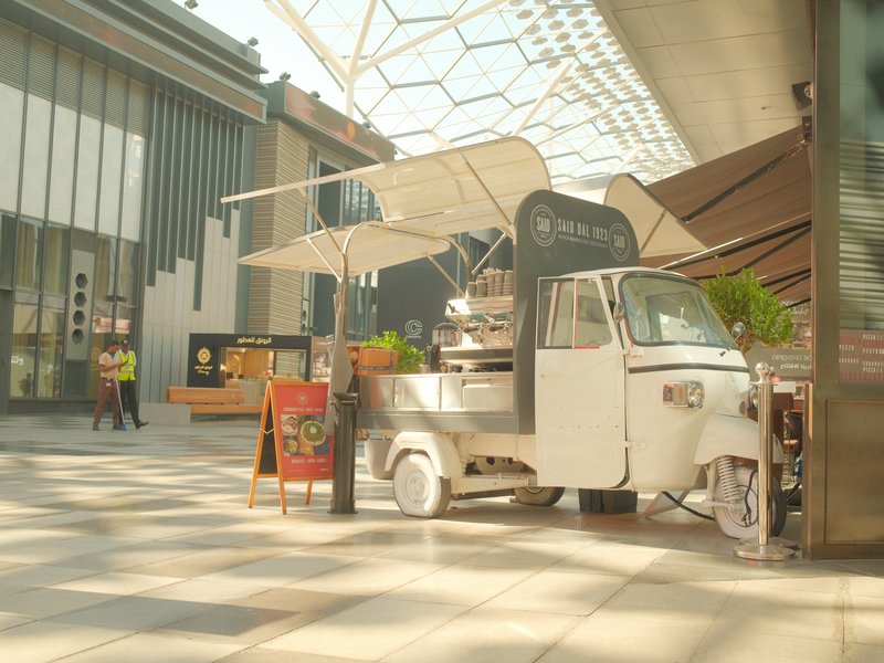 A white tuk-tuk coffee stand inside a modern building with large windows and bright sunlight.