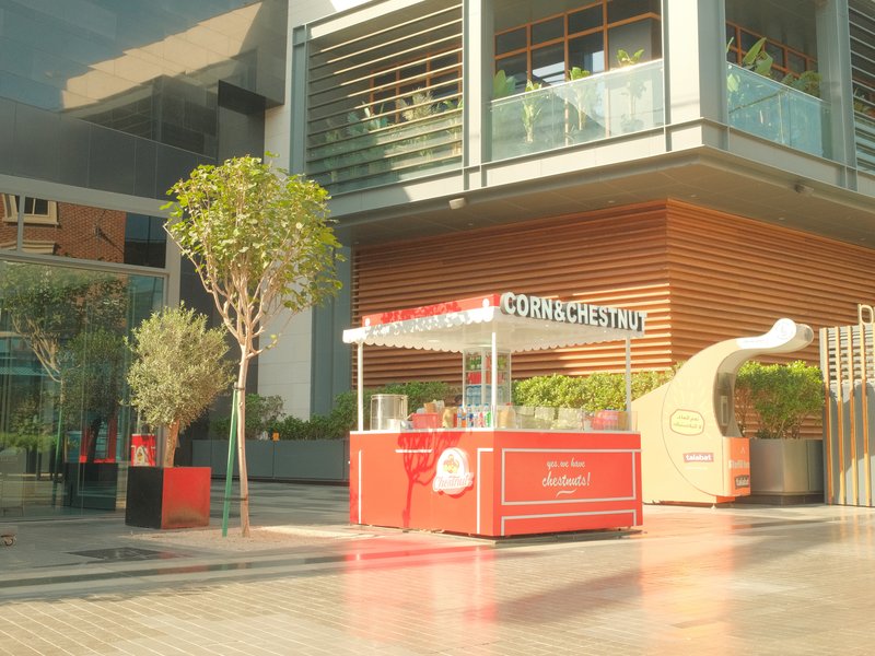 A vibrant outdoor food stall with a red canopy, surrounded by greenery and modern architecture.