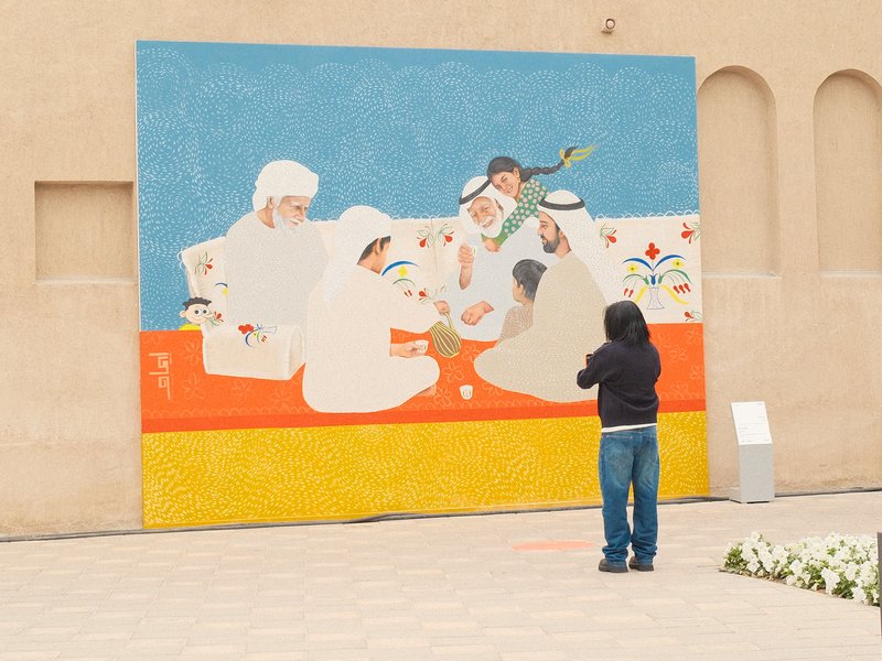 A person photographing a colorful mural depicting people in traditional attire engaged in a cultural activity.