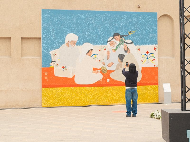A person photographing a colorful mural depicting people sharing a meal in a vibrant outdoor setting.