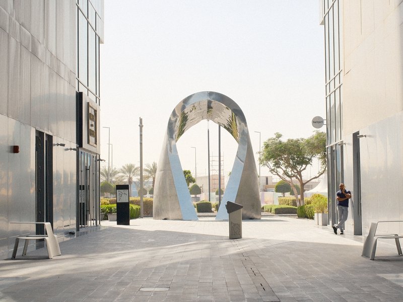 A modern arch sculpture between two sleek buildings in a sunny urban setting.