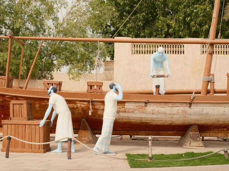Three life-sized figures in traditional attire interacting with a wooden boat in a sunny outdoor setting.