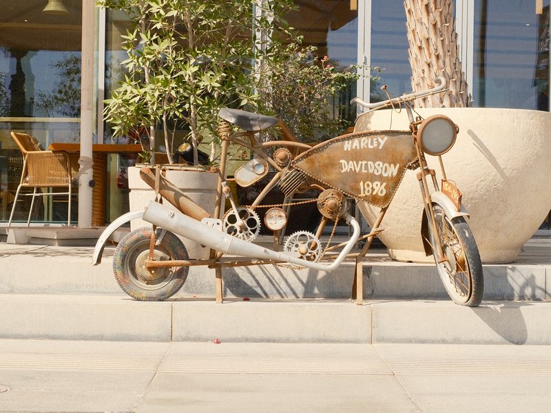 An old, rusty motorcycle displayed outside a modern building with wooden furniture and greenery.