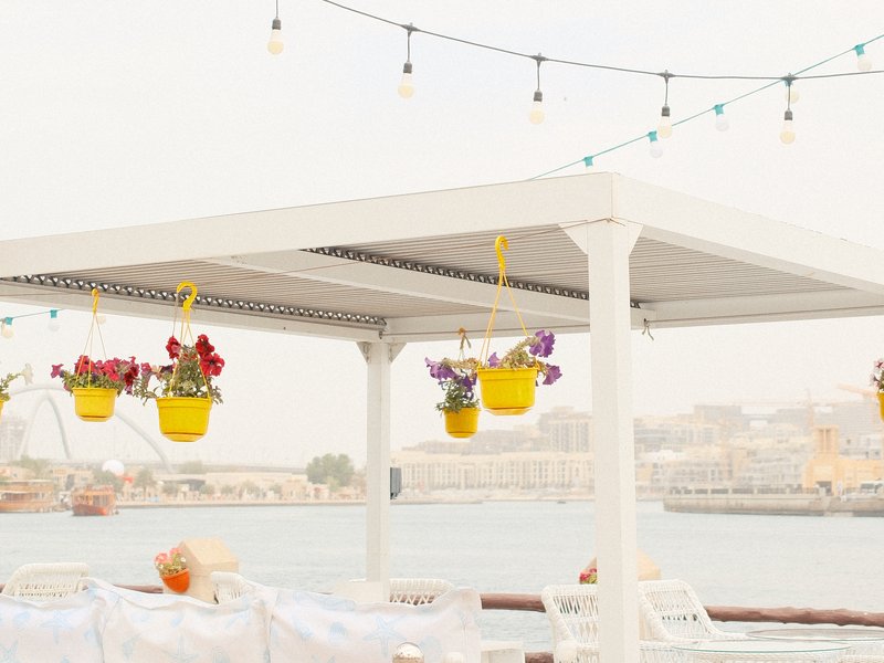 A bright outdoor dining area with hanging flower pots and a view of the water in the background.