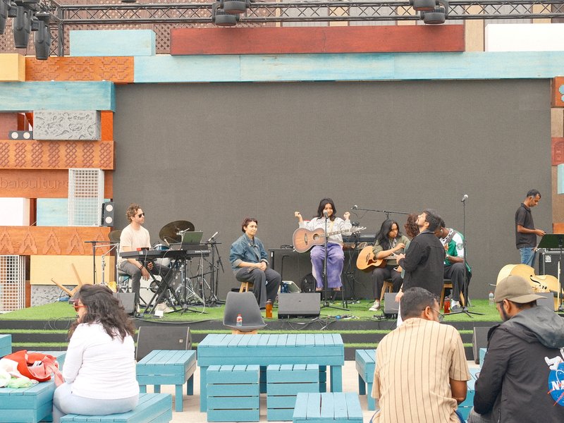 A live band performing on stage with colorful wooden backdrop, audience seated in foreground.