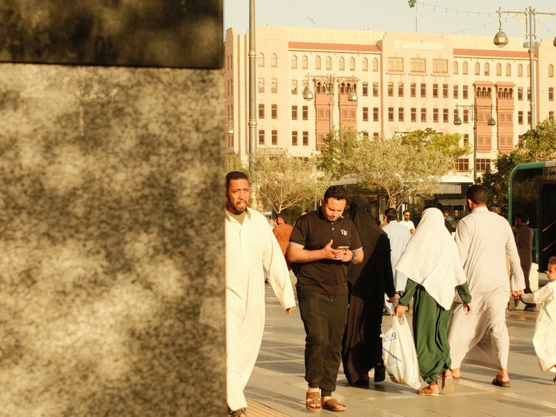 People walking in a busy urban area with a large stone structure in the foreground and buildings in the background.