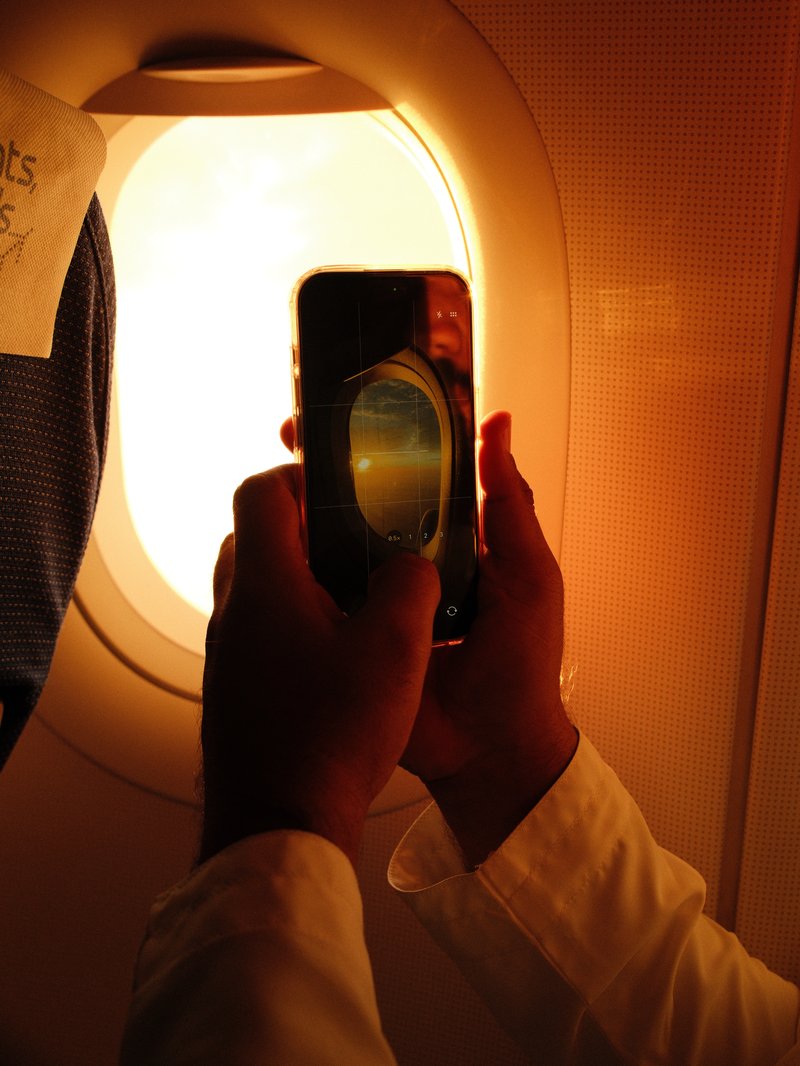 A person holding a smartphone up to an airplane window, capturing the warm glow of sunlight.
