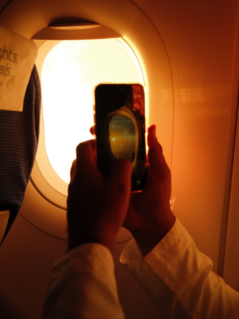 A person holding a smartphone up to an airplane window, capturing a sunset view.