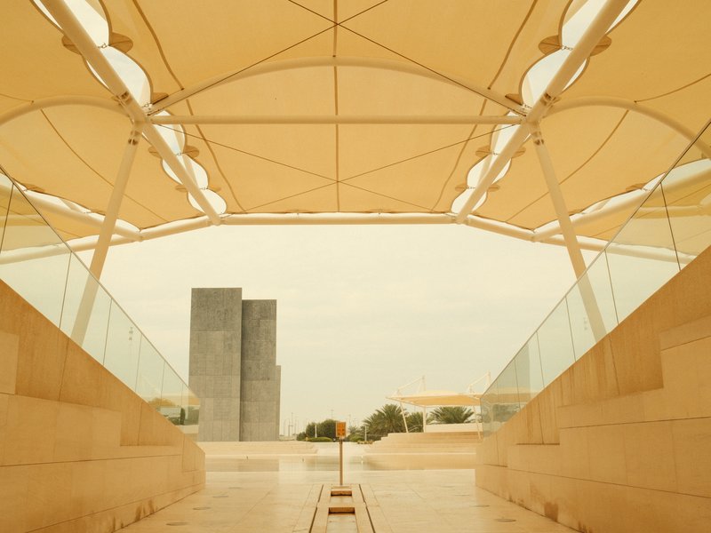 A modern architectural structure with a large canopy and two tall concrete buildings in the background under a cloudy sky.