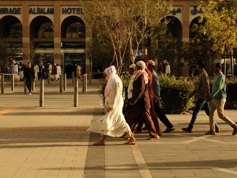 Group of people walking in a city plaza with trees and buildings in the background.