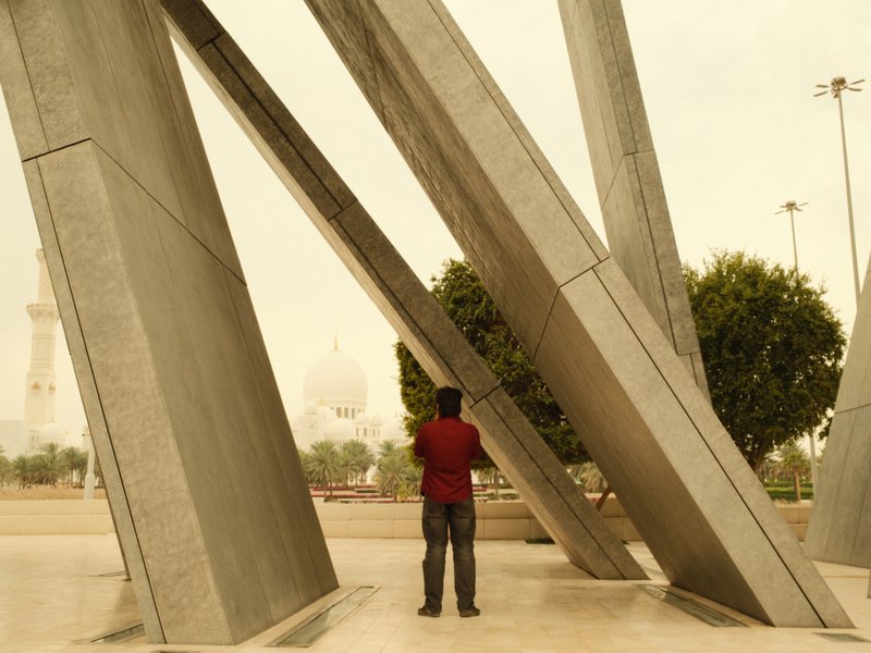 A person in a red shirt stands near large metallic structures in a park, with a dome in the background.