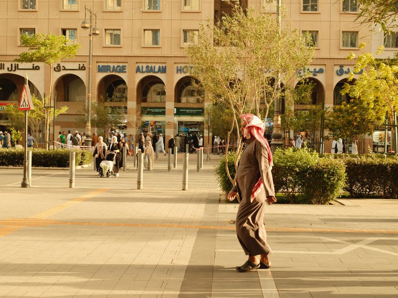 A man in traditional attire walking through a bustling urban plaza with greenery and people around.
