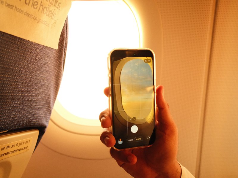 A hand holding a smartphone capturing a view of an airplane window with sunlight streaming through.