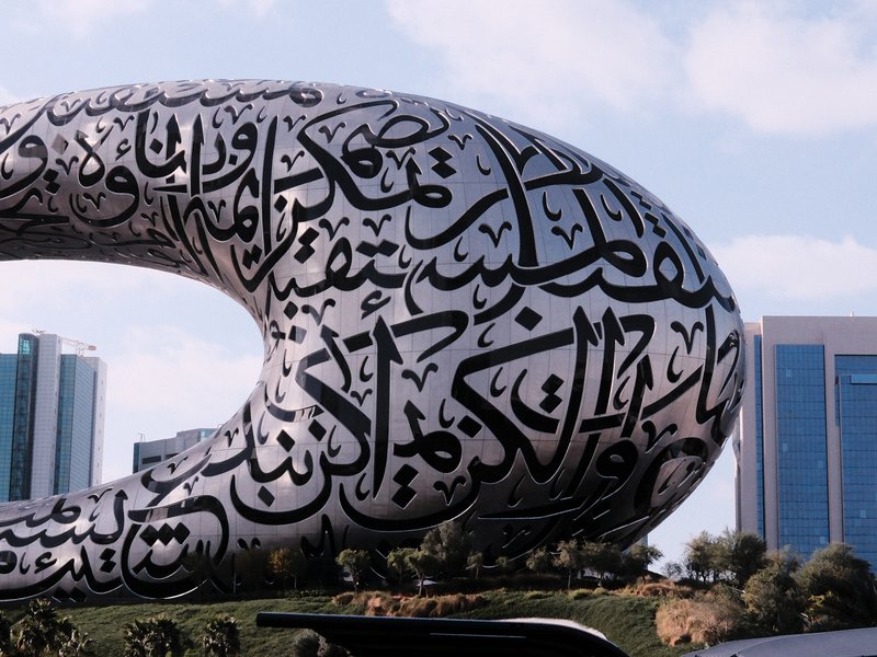A large, modern sculpture covered in Arabic calligraphy, set against a backdrop of buildings.